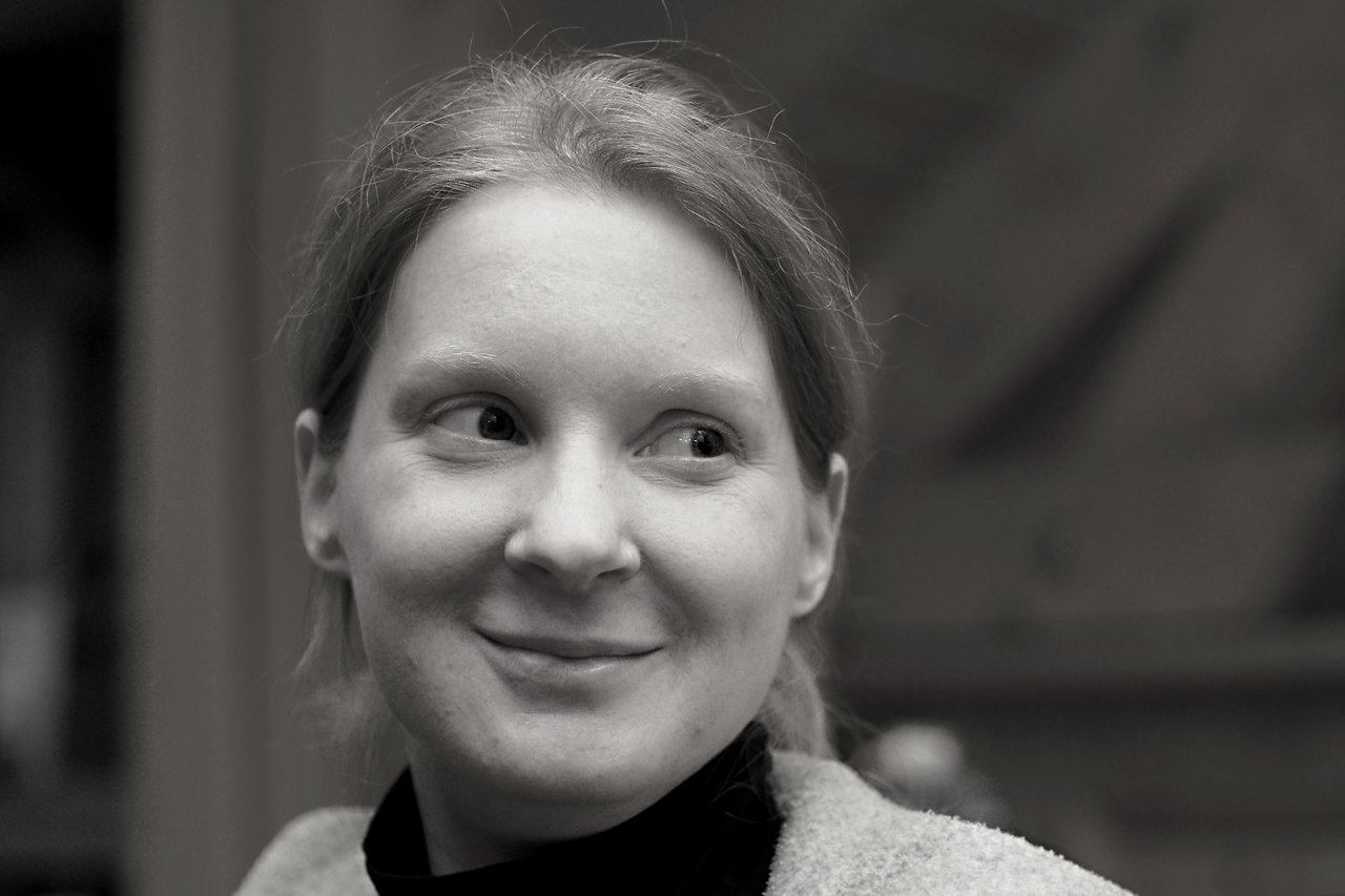 A woman with light skin and tied-back hair smiles slightly while looking to the side in a black-and-white portrait.