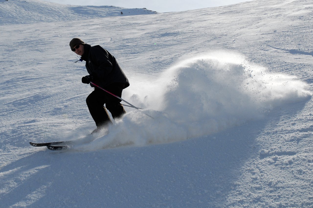 A skier in dark clothing makes a sharp turn, kicking up a spray of snow on a snowy slope.