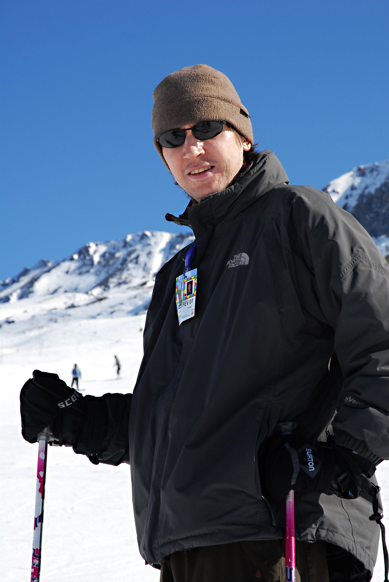 A person wearing a winter jacket, hat, and sunglasses holds ski poles on a snowy mountain slope.