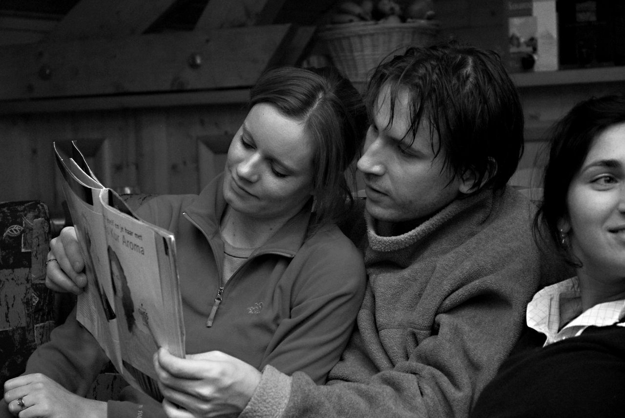 A man and woman sit closely together on a couch, reading a magazine, while another woman smiles nearby.