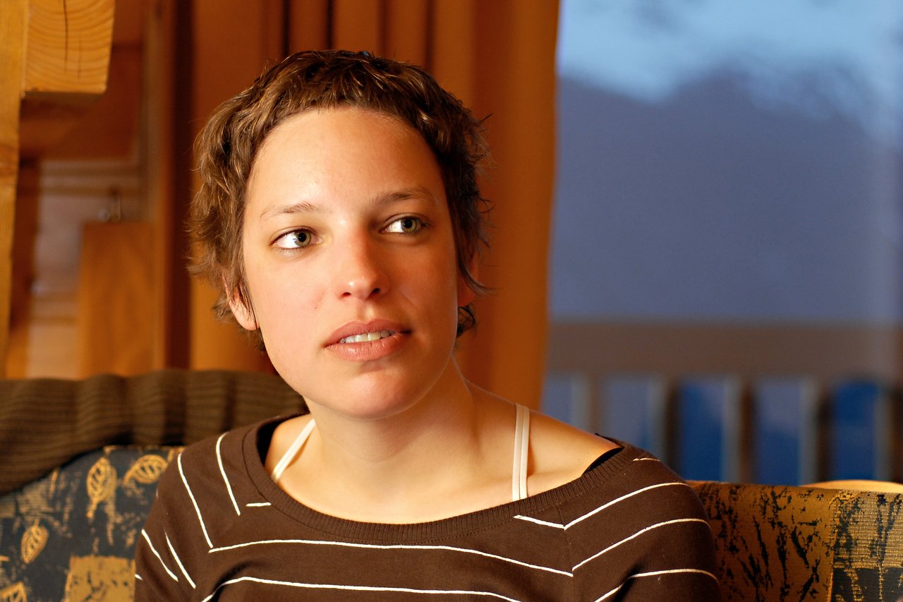 A woman with short brown hair sits indoors, looking slightly to the side with a neutral expression.