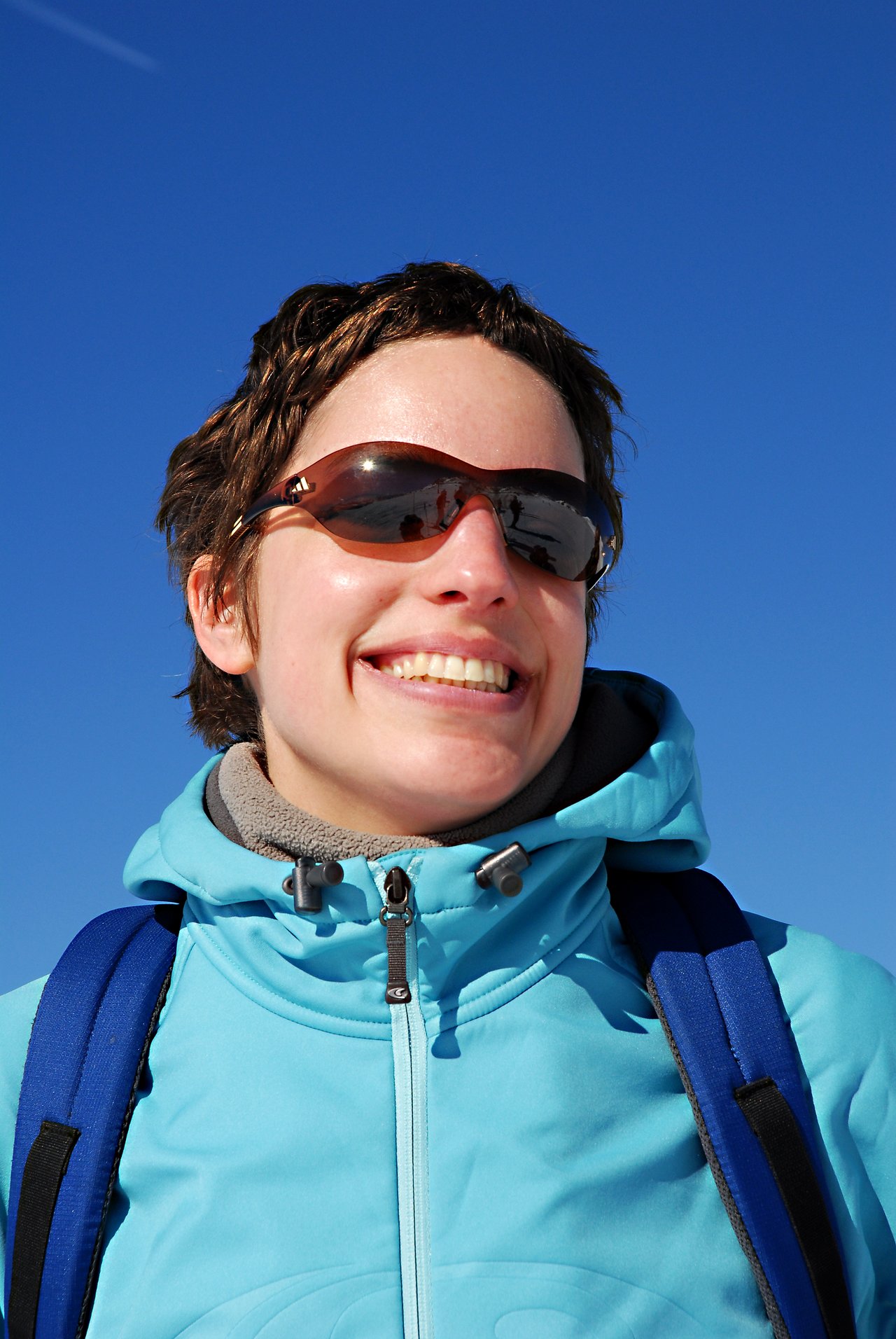 A person wearing sunglasses, a blue jacket, and a backpack smiles outdoors under a clear blue sky.