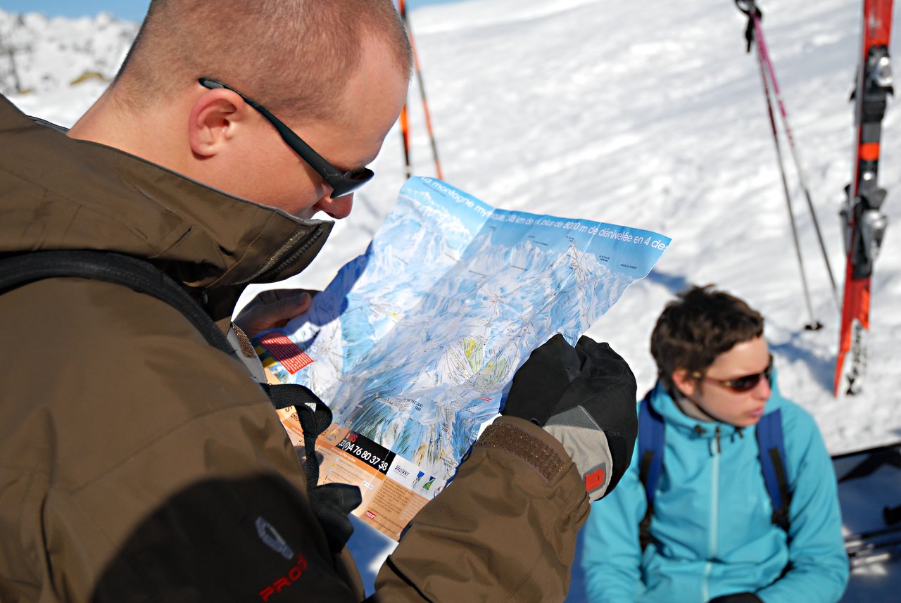 A person in a brown jacket and sunglasses studies a ski map, while another person sits nearby in the snow.