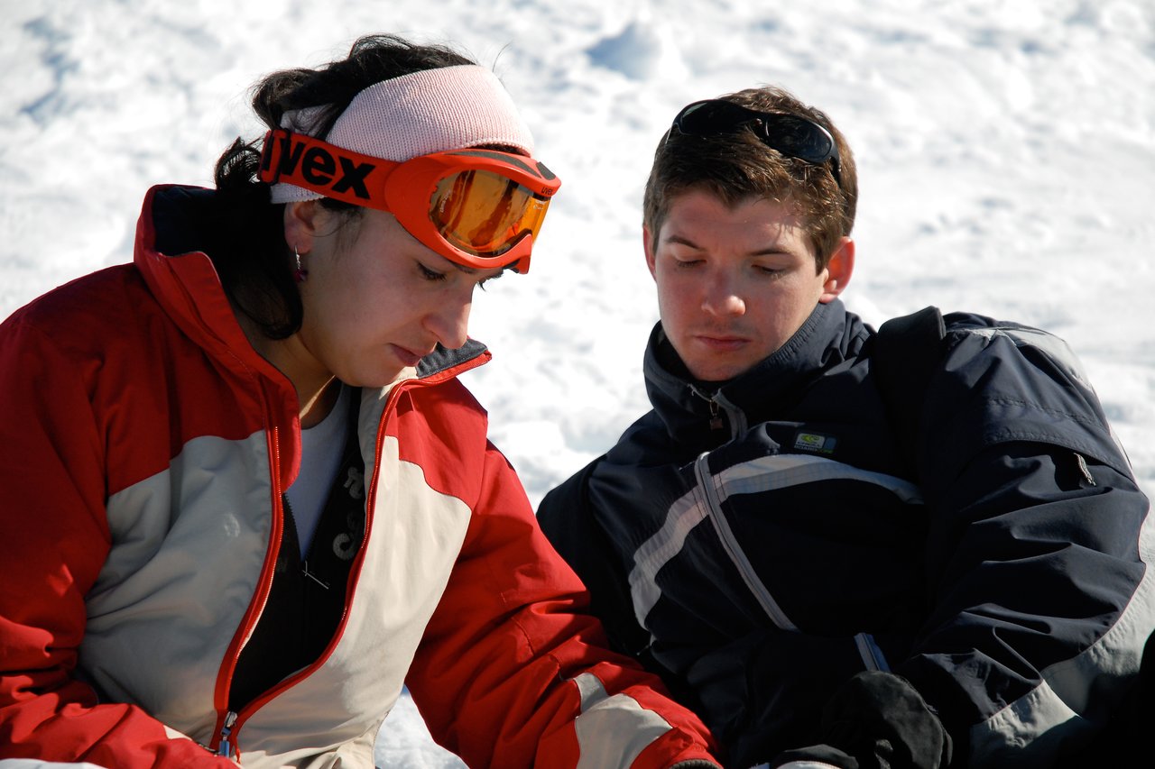 Two people in winter gear sit on the snow, one looking down with a pained expression.