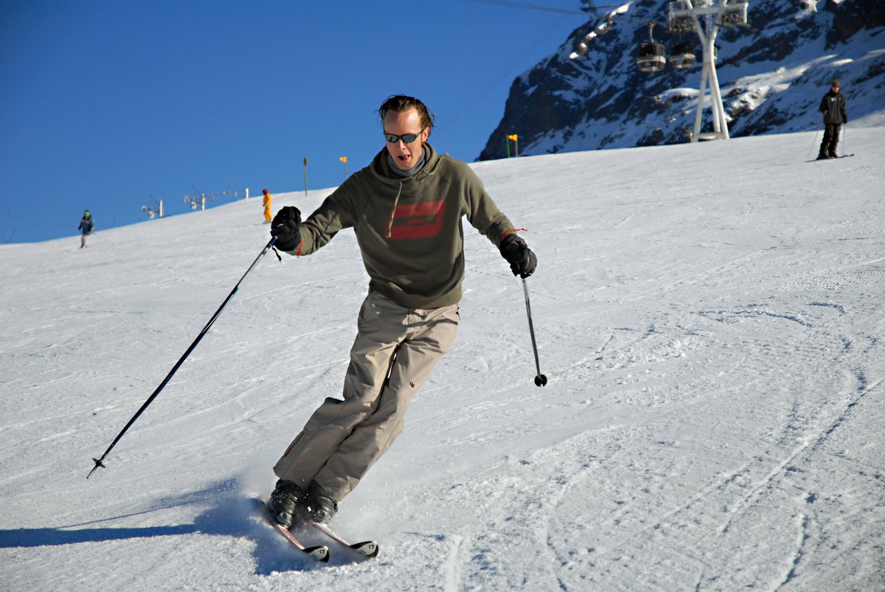 A skier in a green hoodie and beige pants makes a sharp turn on a snowy slope, holding ski poles.