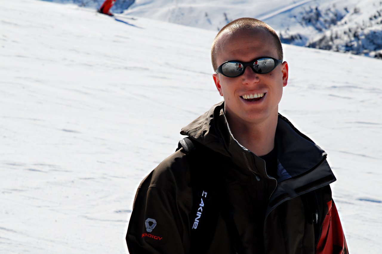 A person wearing sunglasses and a winter jacket smiles while standing on a snowy mountain slope.