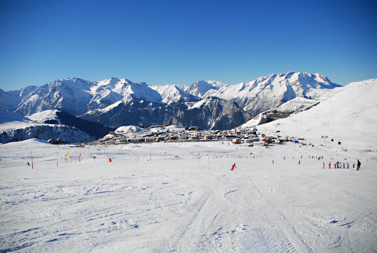Skiers and snowboarders on a snowy slope with a mountain village and snow-covered peaks in the background.