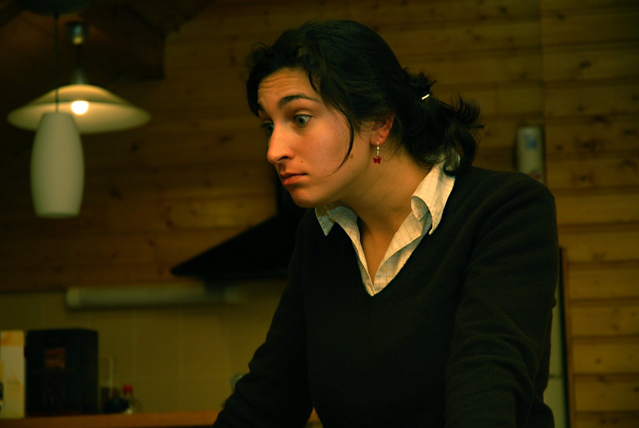 A woman with dark hair and red earrings leans forward, looking intently at something with a focused expression.