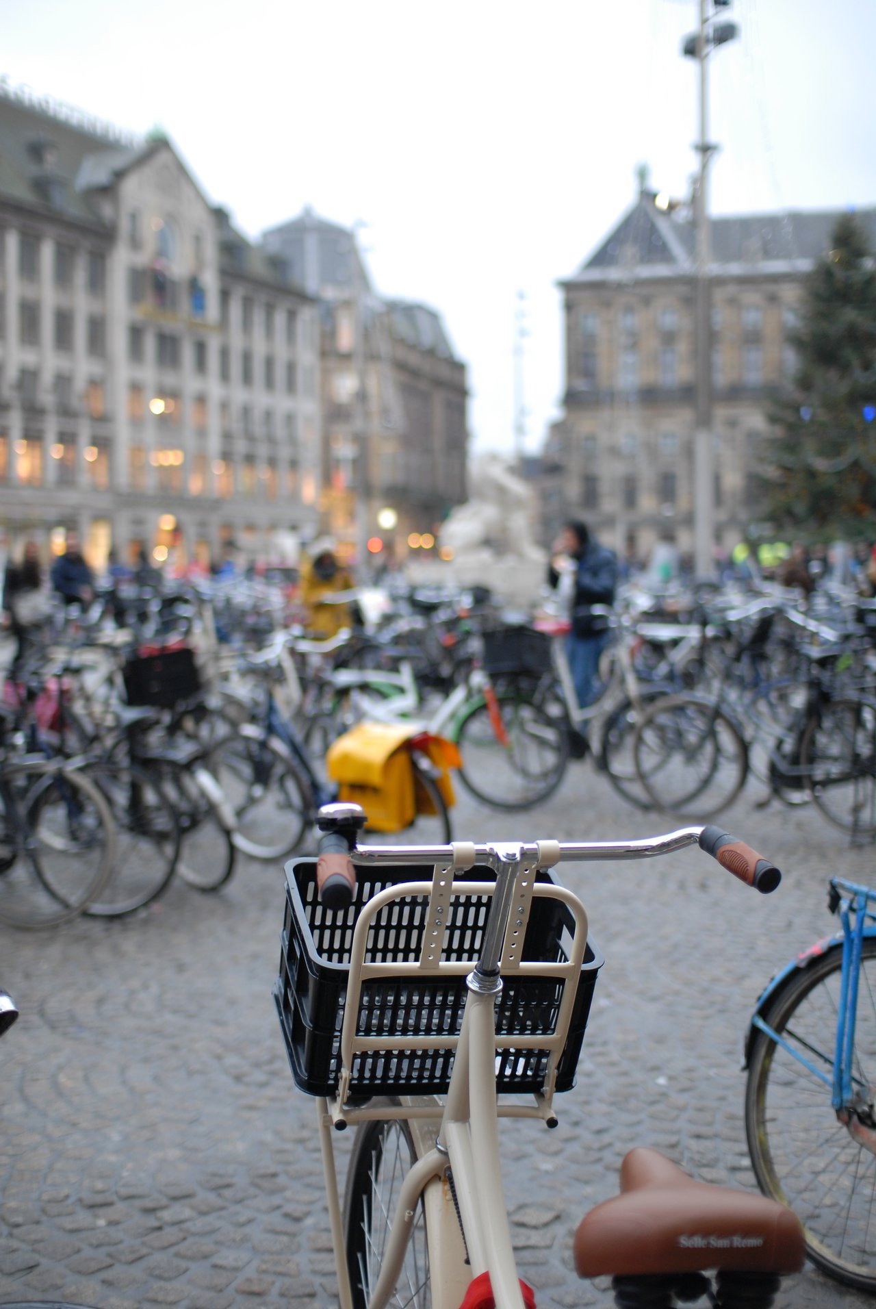 A beige bicycle with a black basket in the foreground, surrounded by many parked bikes in a busy square.