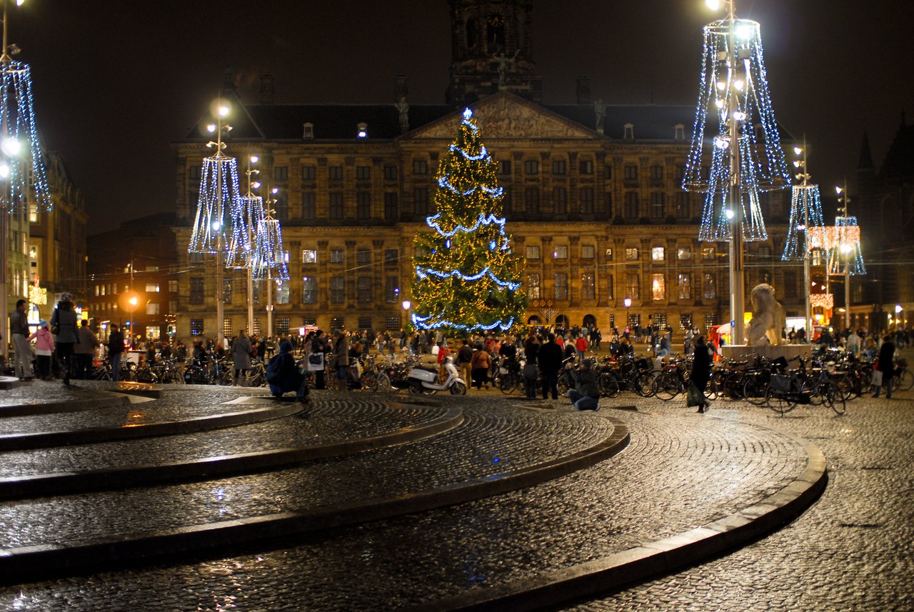 A large Christmas tree with lights stands in a busy square at night, surrounded by people and bicycles.