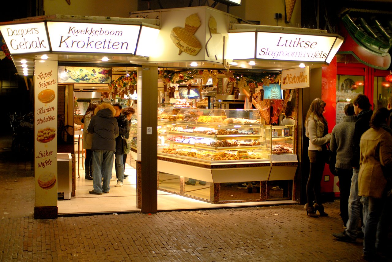 A bakery stand with people ordering and browsing pastries, including kroketten and waffles, on a busy evening street.