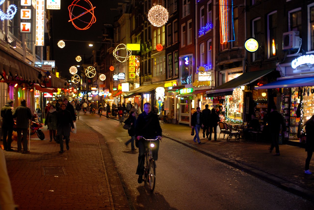 A person rides a bicycle on a busy, well-lit street at night, surrounded by pedestrians and shops.
