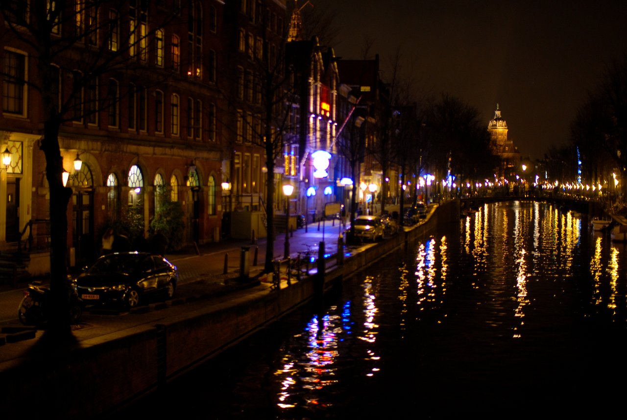 A canal at night in Amsterdam, with streetlights and neon signs reflecting on the water.