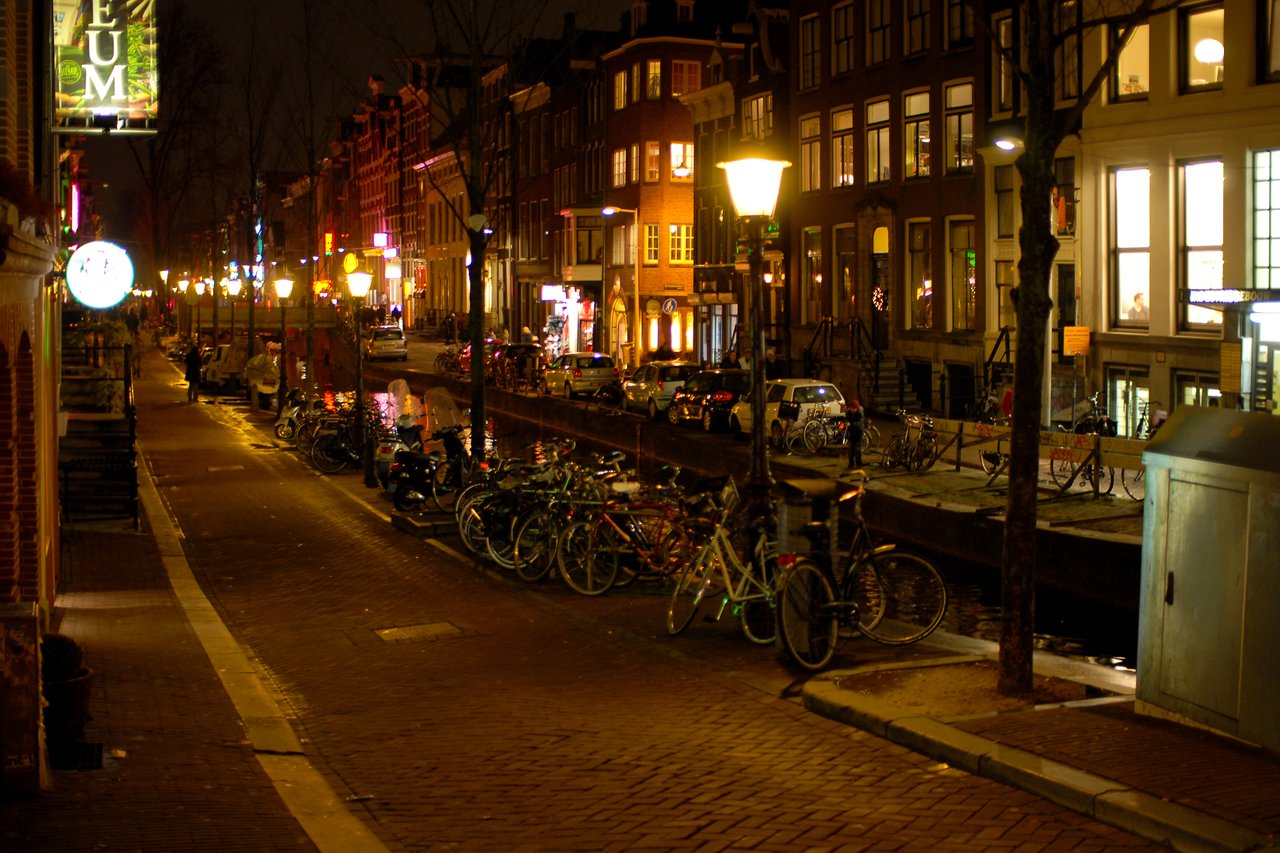 A canal-side street in Amsterdam at night, with bicycles parked along the railing and illuminated buildings in view.