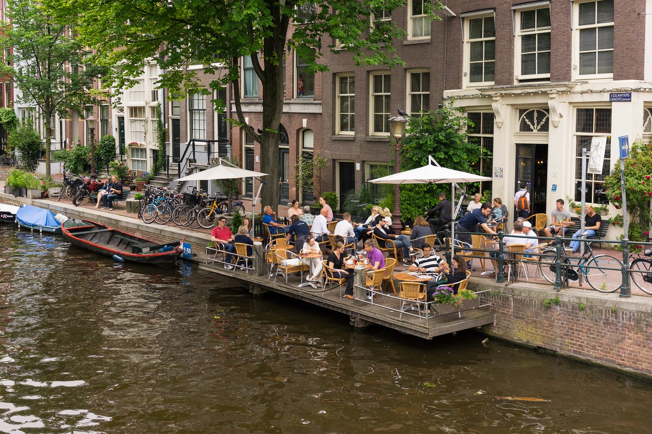 People sitting at an outdoor café by a canal in Amsterdam, enjoying food and drinks under umbrellas.