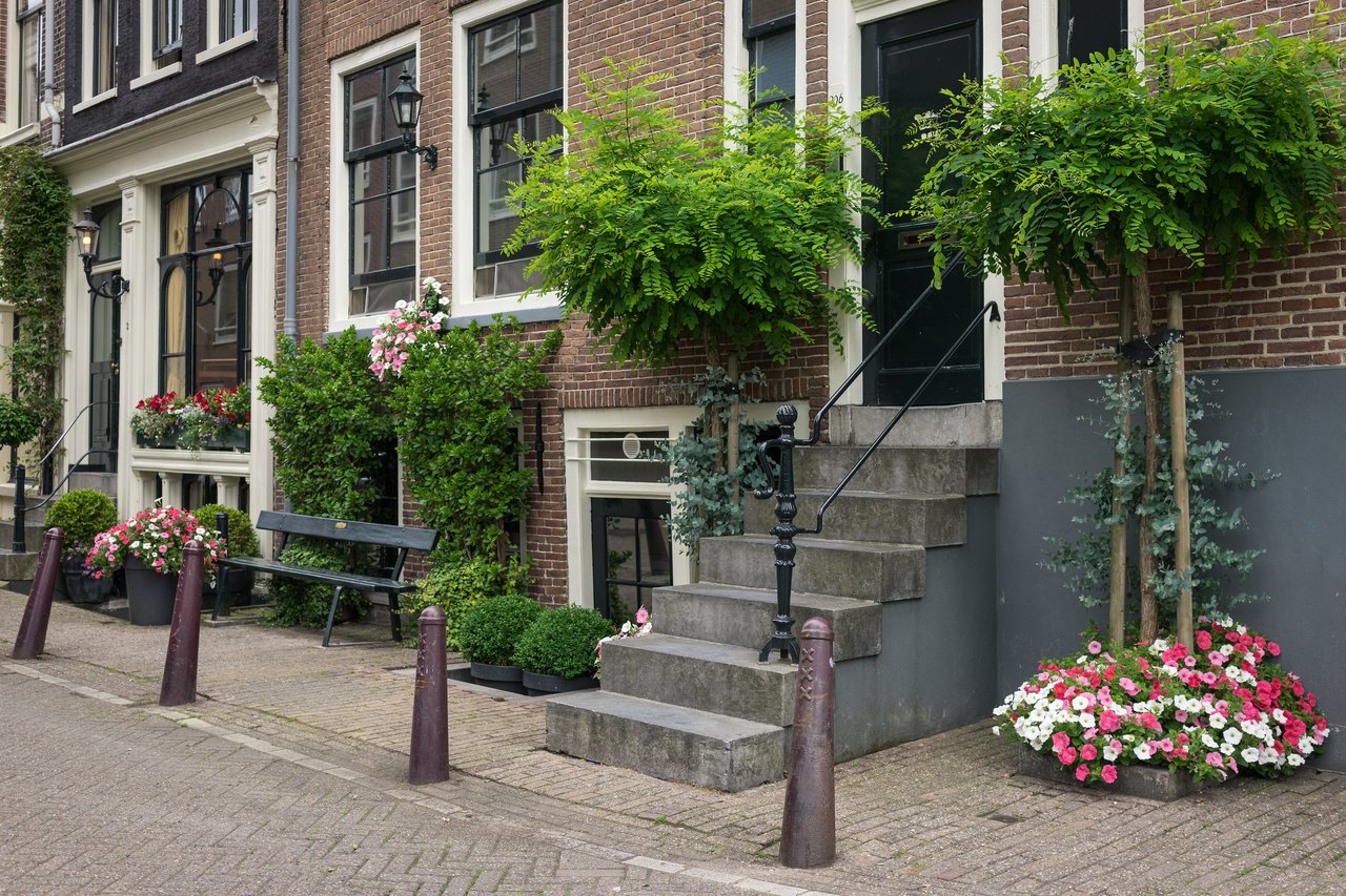 A brick building in Amsterdam with a black door, stone steps, and colorful flowers in planters along the sidewalk.