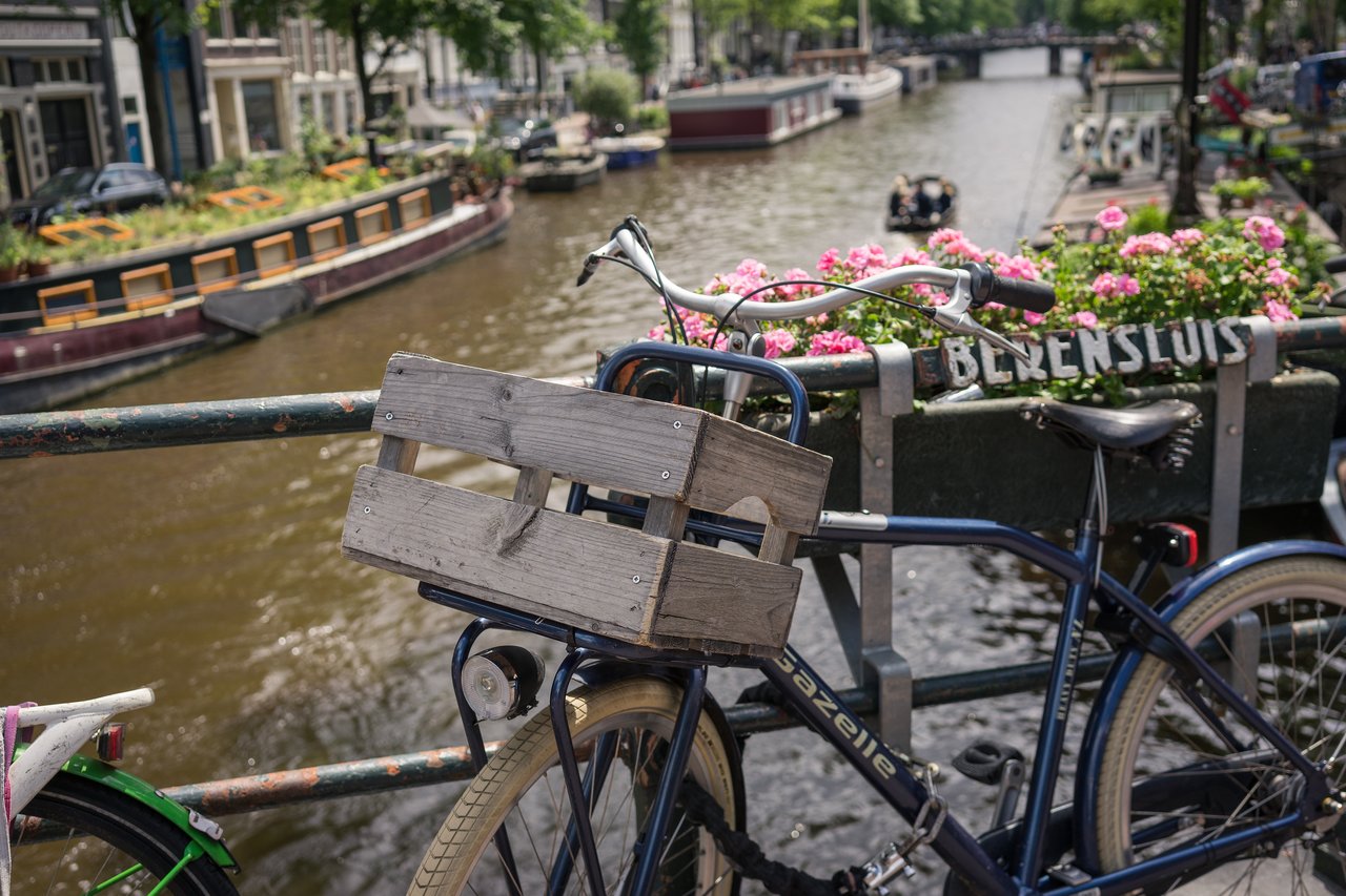 A bicycle with a wooden crate is parked on a bridge overlooking an Amsterdam canal with boats and flowers.