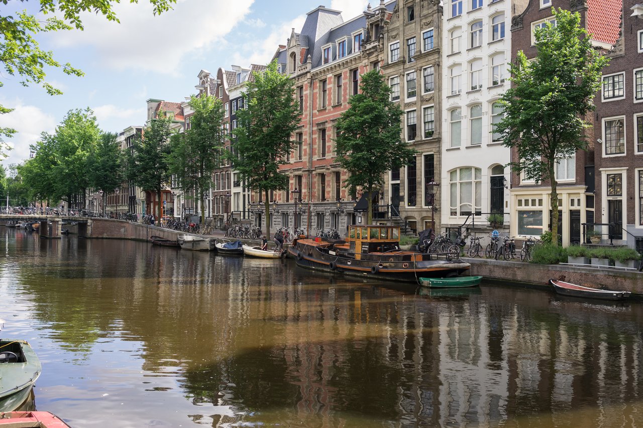 A canal in Amsterdam with boats docked along the water, lined with tall, narrow buildings and trees.