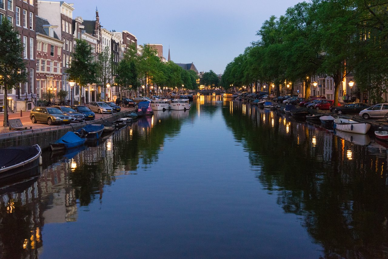 A calm canal in Amsterdam at night, with boats, parked cars, and streetlights reflecting on the water.