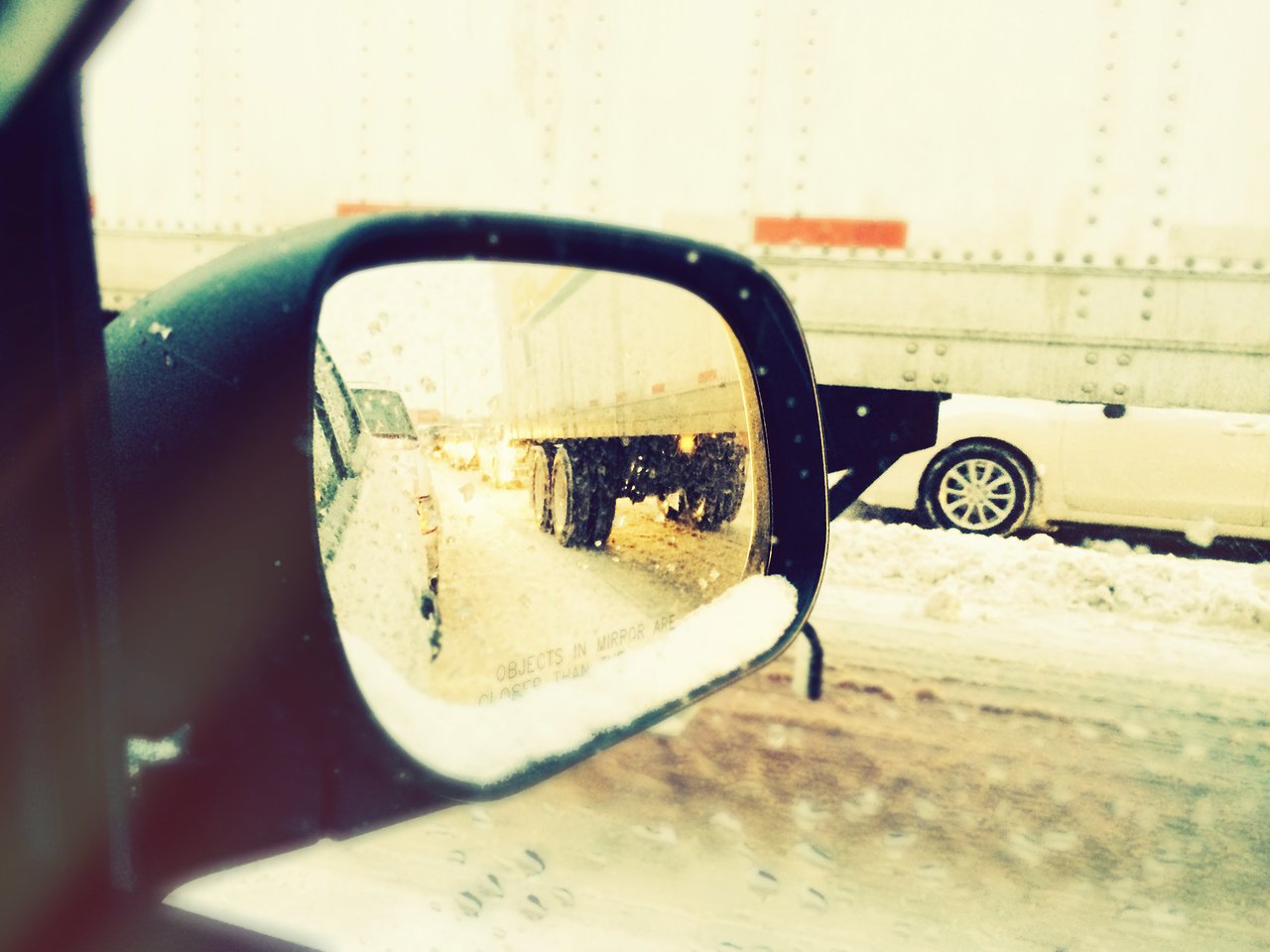 A car's side mirror reflects a snowy traffic jam with trucks and cars on a slushy road.