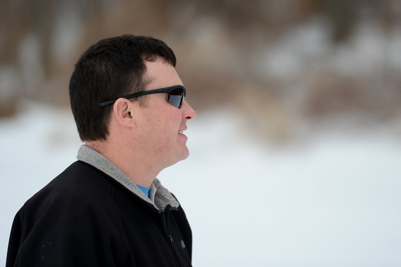 A man wearing sunglasses and a black jacket looks to the side in a snowy outdoor setting.