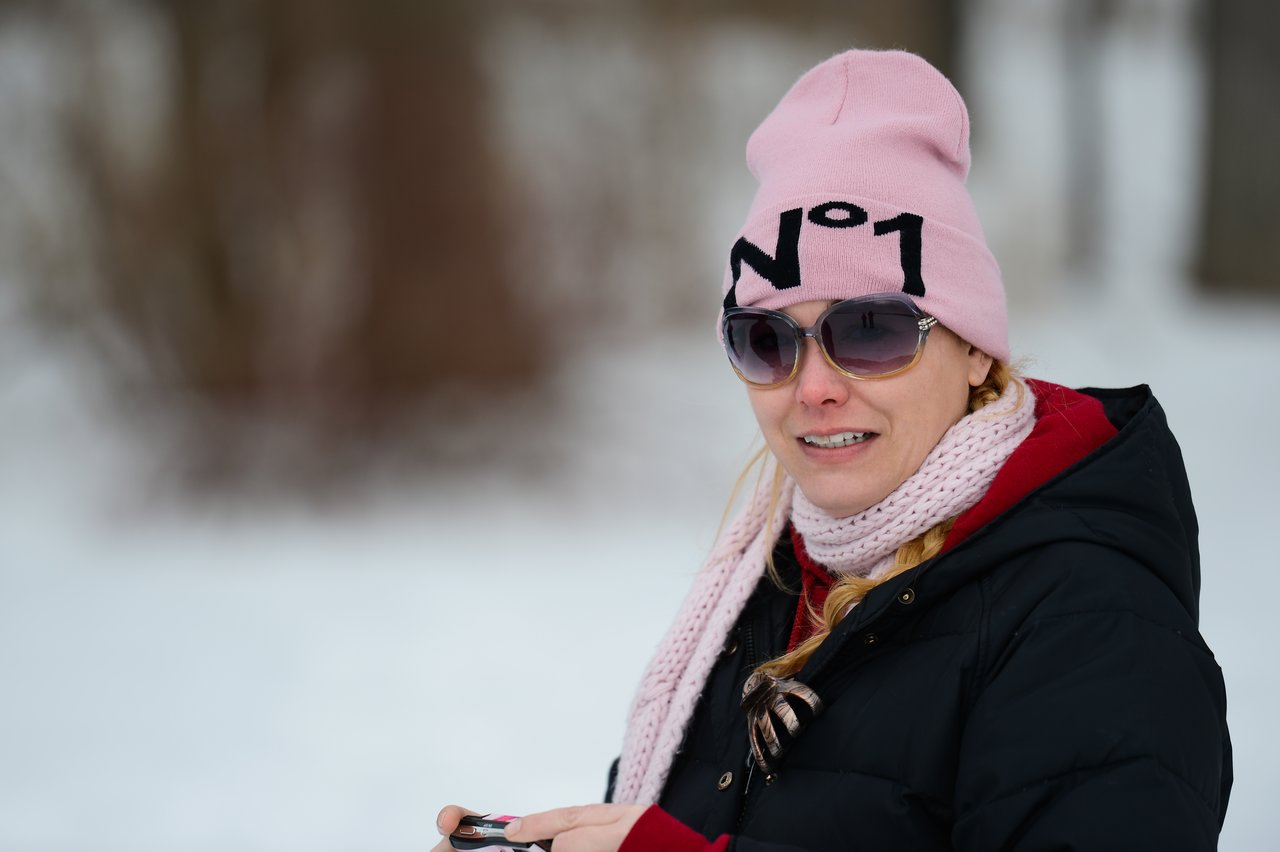 A woman wearing a pink beanie, sunglasses, and a scarf stands outside in the snow, holding a small device.
