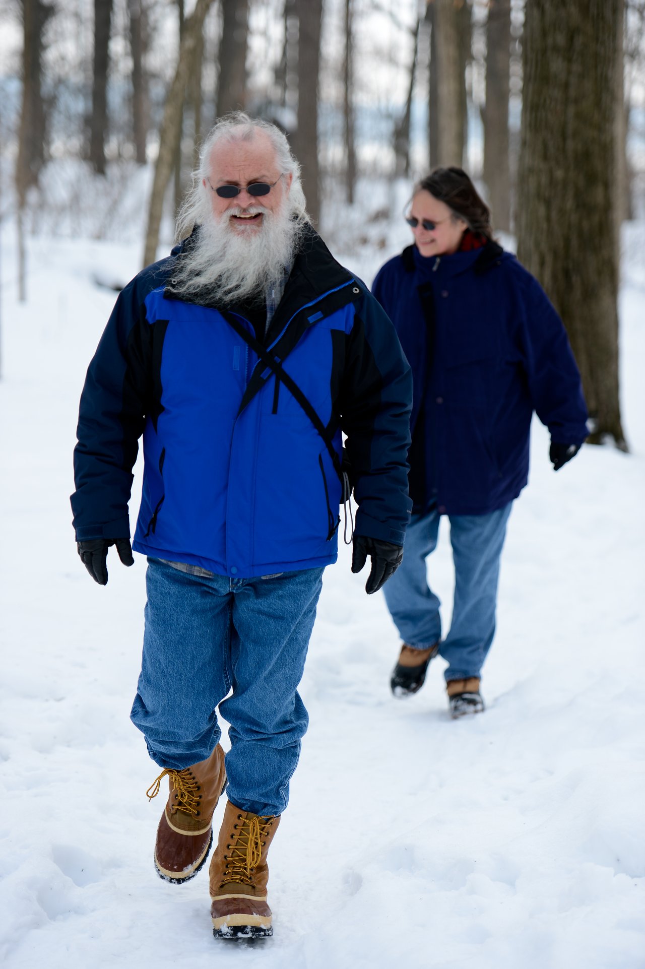 Two people walk through the snow in a wooded area, wearing winter jackets, gloves, and boots.