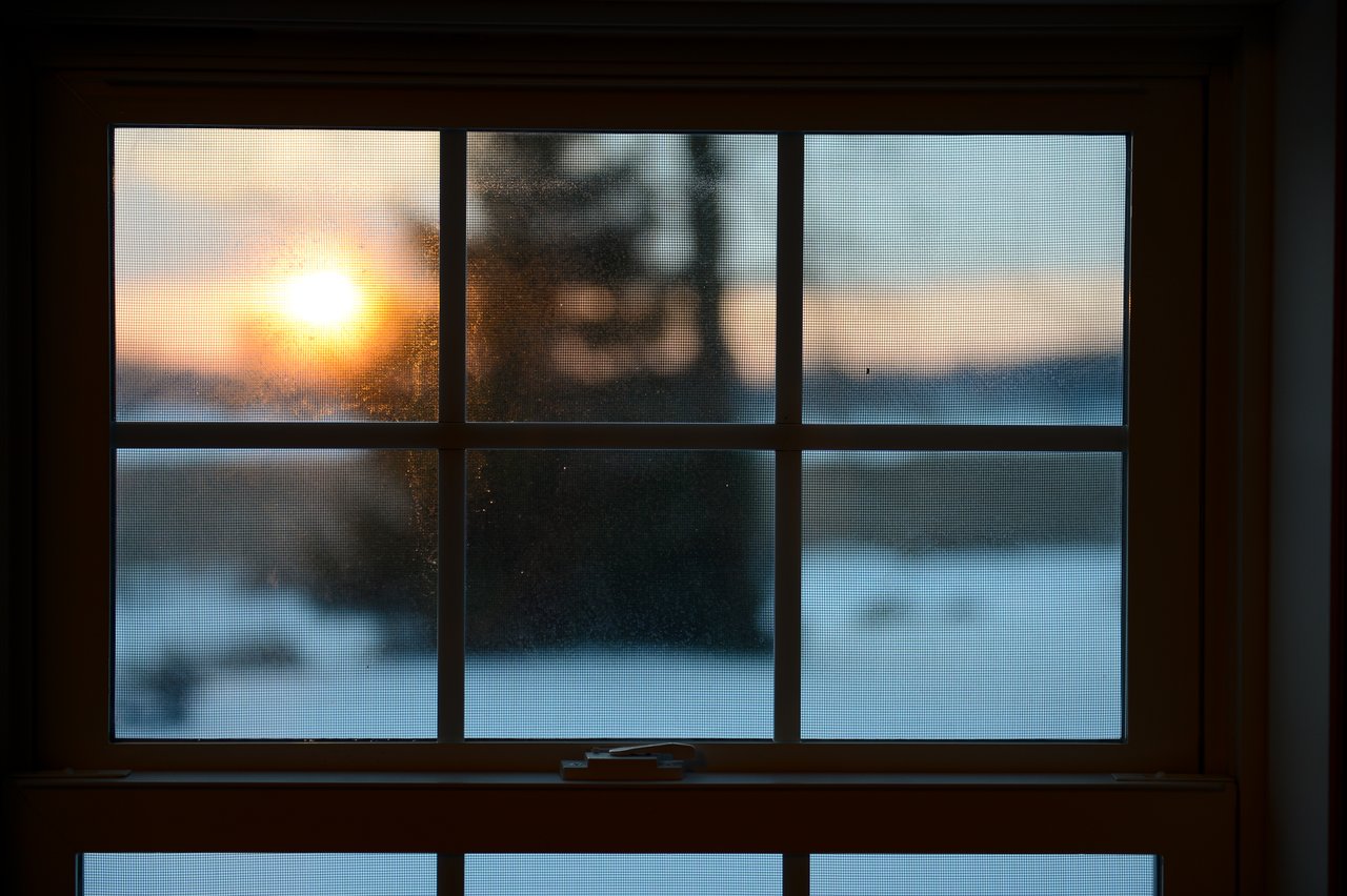 Sunset seen through a window with a screen, overlooking a snowy landscape with a tree in the distance.