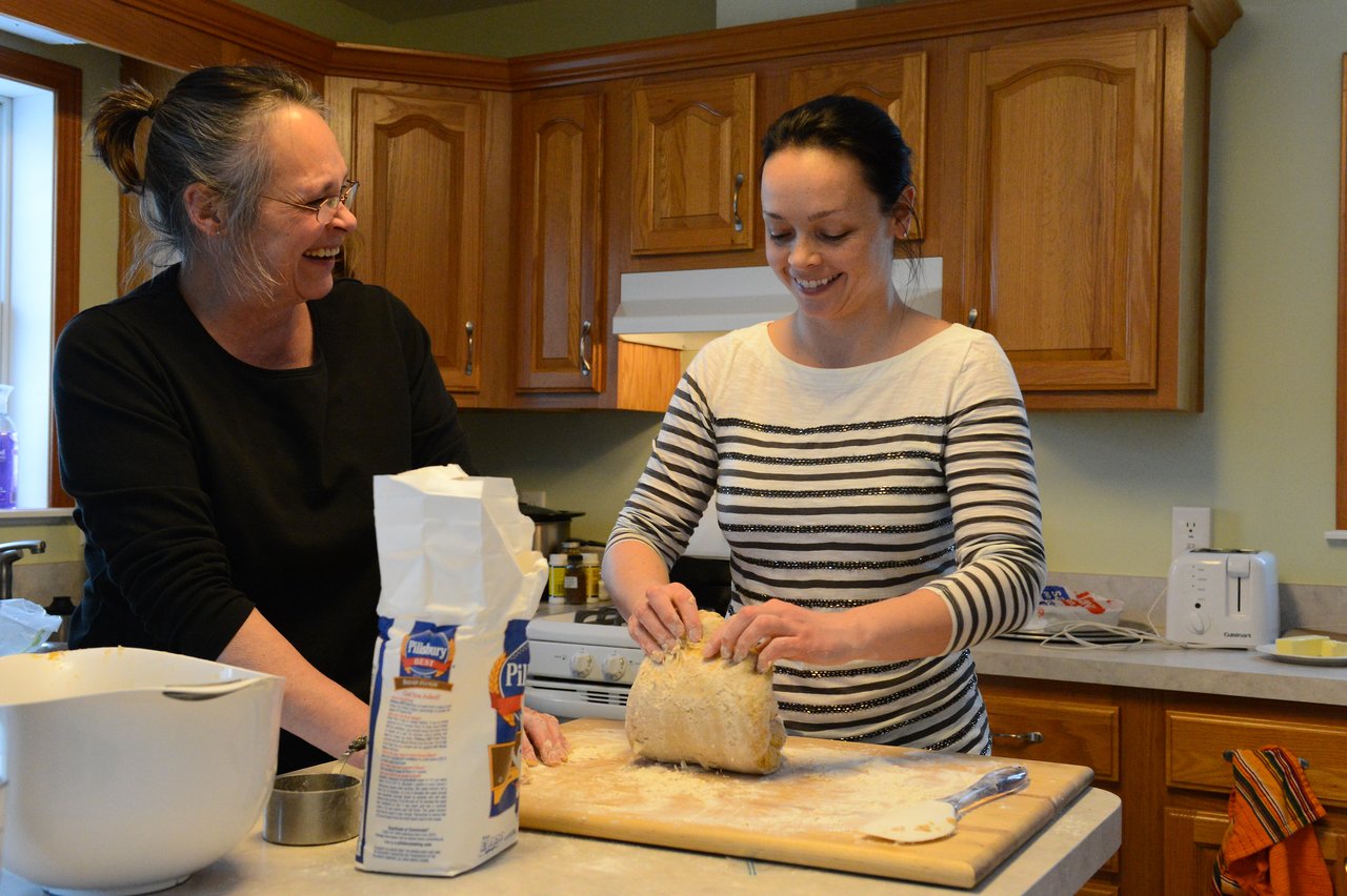 Two women smile while kneading dough on a wooden board in a kitchen with baking ingredients nearby.