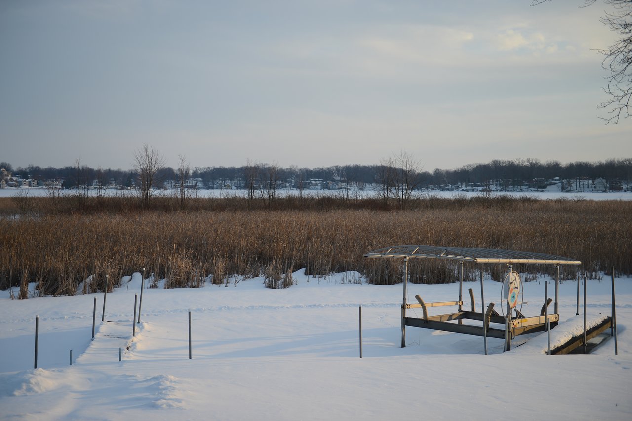A snow-covered dock with a metal canopy stands near a frozen lake, surrounded by tall, dry grass.