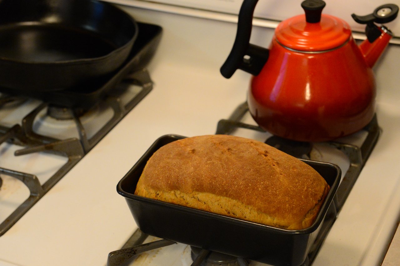 A freshly baked loaf of bread sits in a black pan on a stovetop near a red kettle.