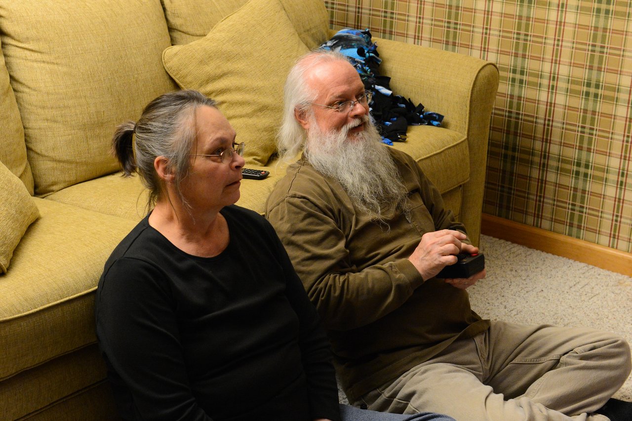 An older man and woman sit on the floor, watching something.