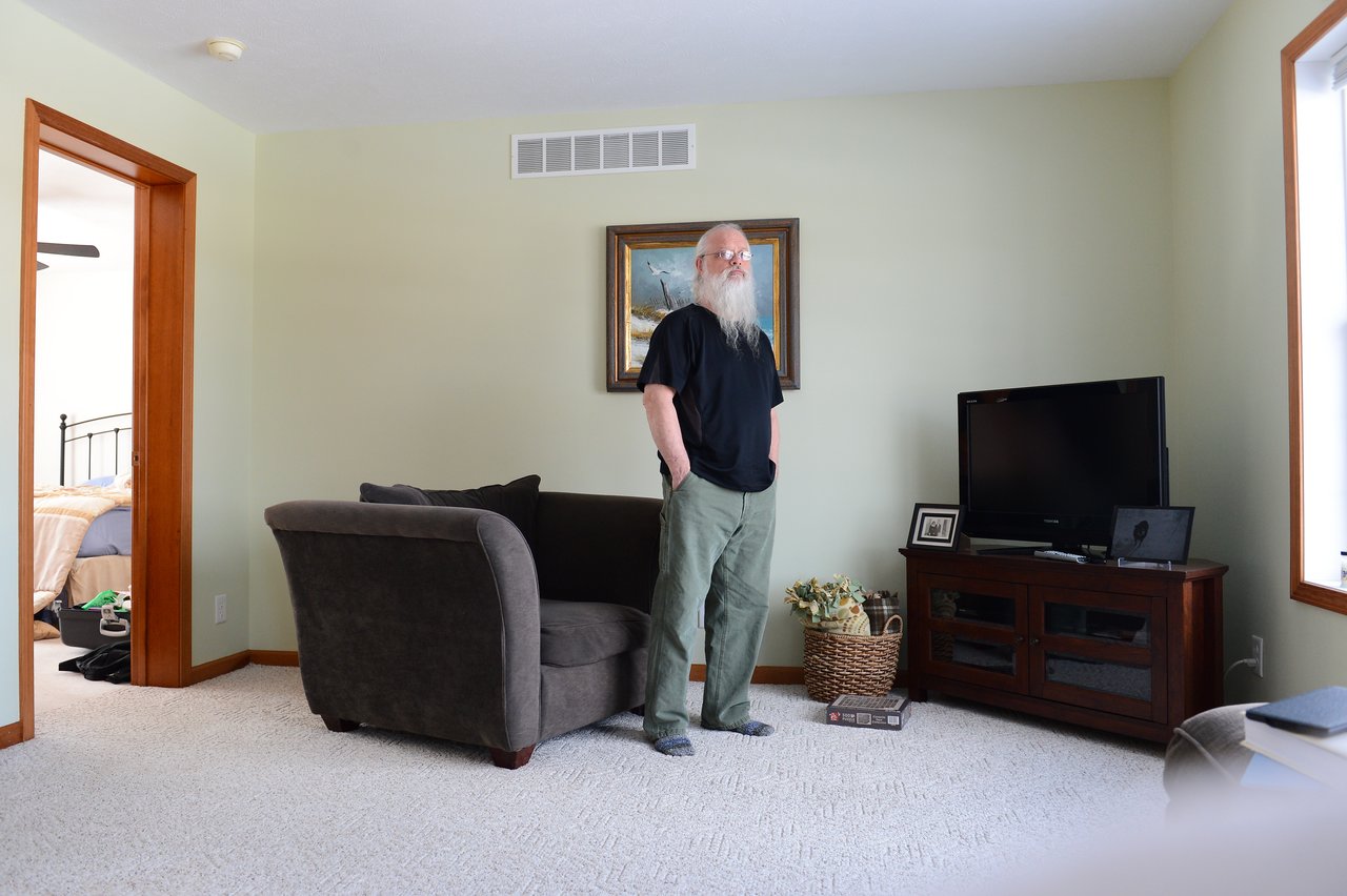 An older man with a long white beard stands in a living room near a couch, looking toward the camera.