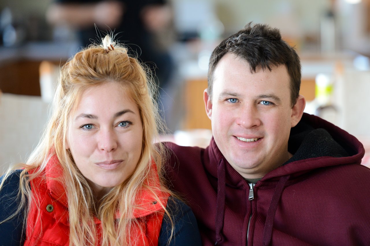 A man and a woman sit closely together, both smiling at the camera in a well-lit indoor setting.