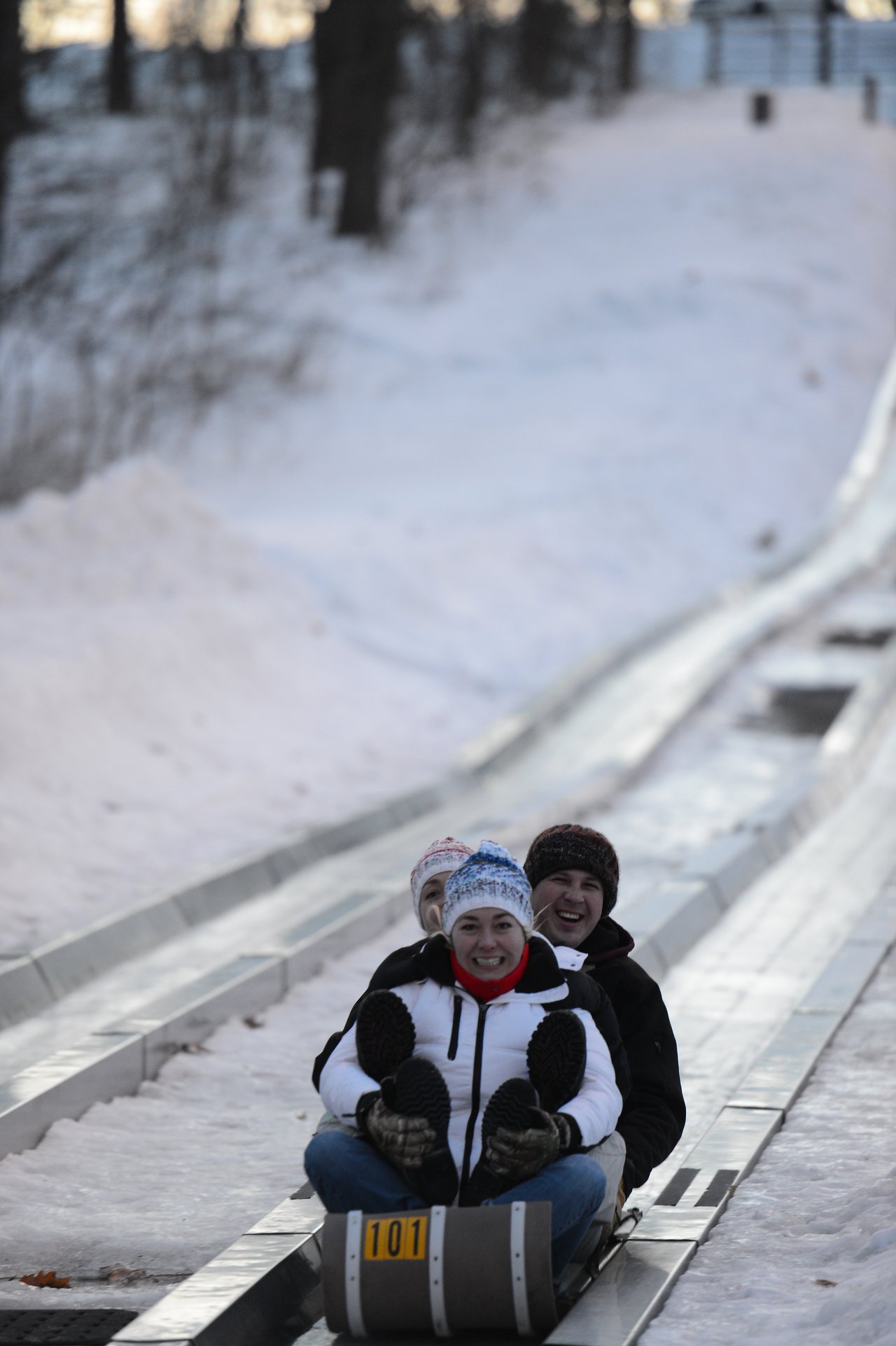 Three people in winter clothing ride a sled down an icy track, smiling and enjoying the snowy outdoor activity.