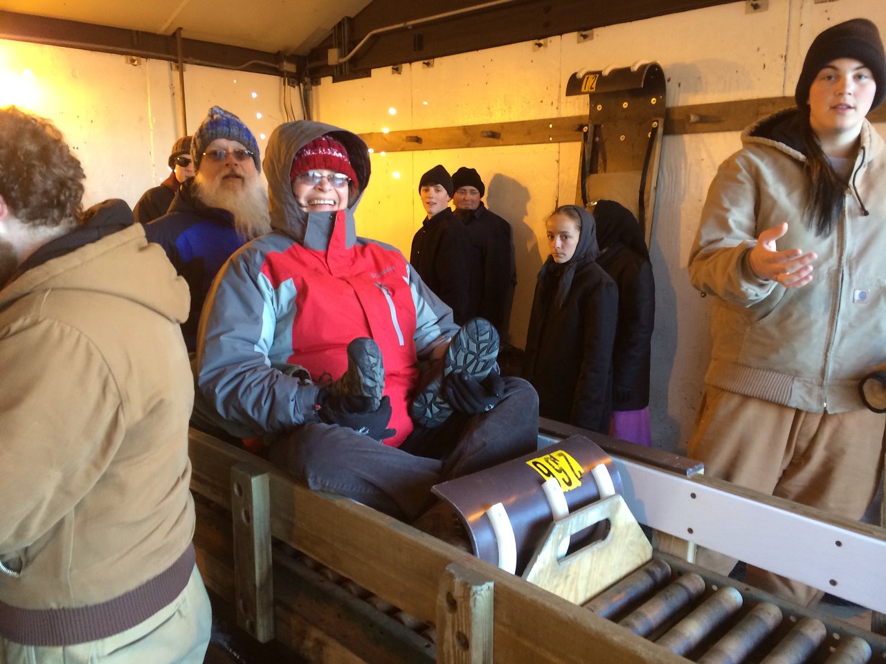 A person in a red and gray jacket sits on a wooden sled, smiling, while others stand nearby.
