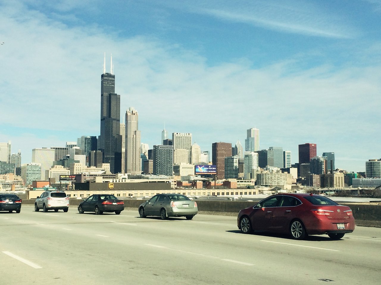 Cars driving on a highway with a city skyline in the background under a clear sky.