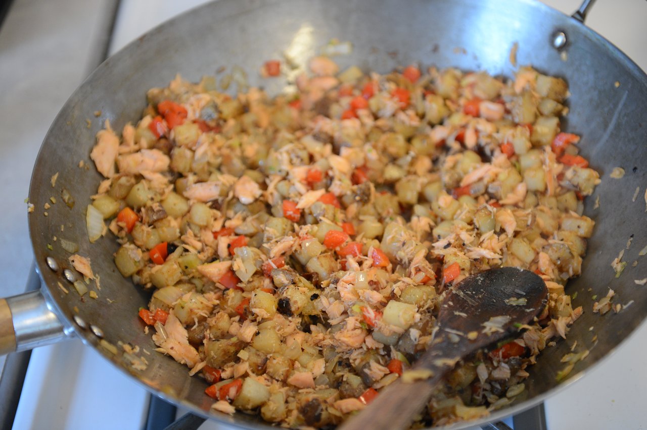 A pan with diced potatoes, red bell peppers, and flaked fish being cooked, with a wooden spoon stirring.