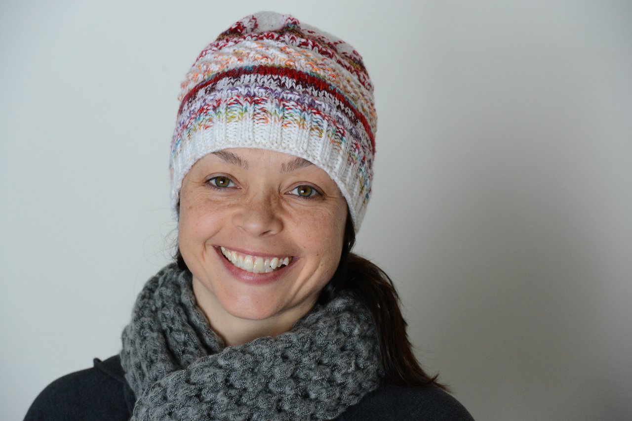 A woman wearing a colorful knit hat and gray scarf smiles at the camera against a plain background.