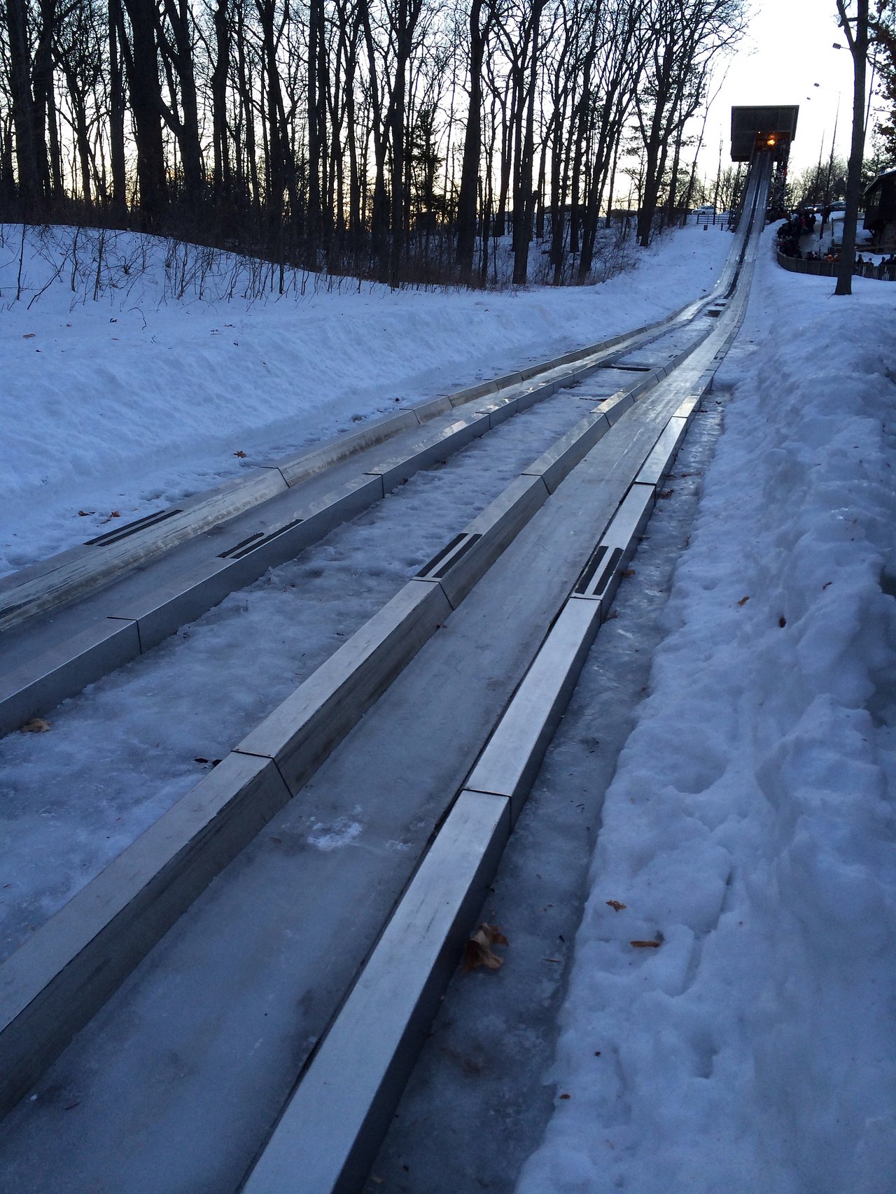 A snowy luge track with metal rails extends downhill, surrounded by trees, leading to a platform at the top.