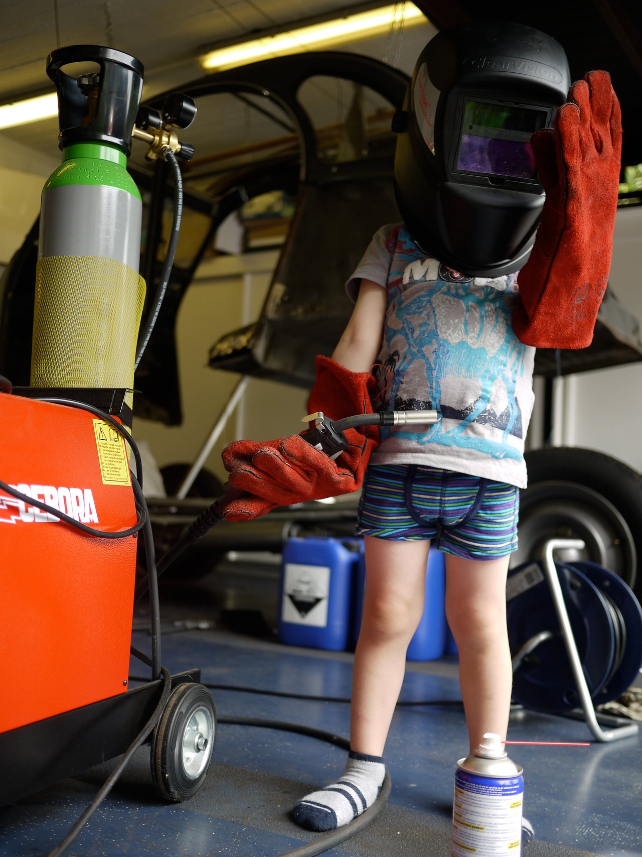 A child wearing a welding helmet and gloves holds a welding torch in a garage or workshop setting.