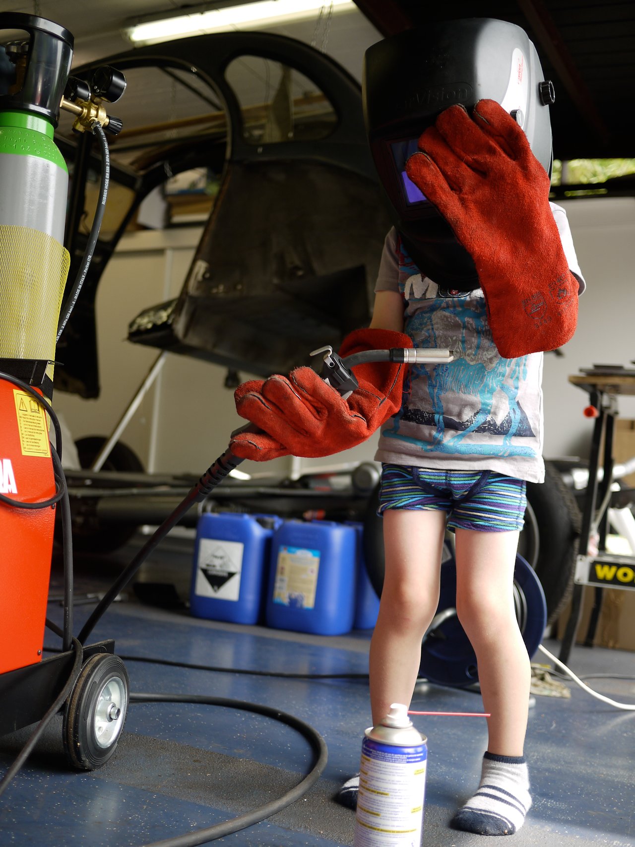 A child wearing a welding helmet and gloves holds a welding torch in a workshop with equipment and tools.