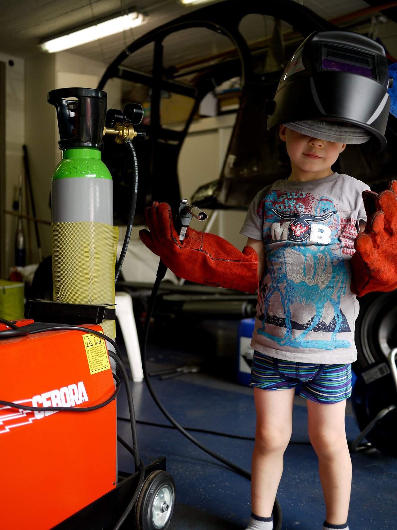 A child wearing a welding helmet and gloves holds welding equipment in a workshop, standing next to a gas cylinder.