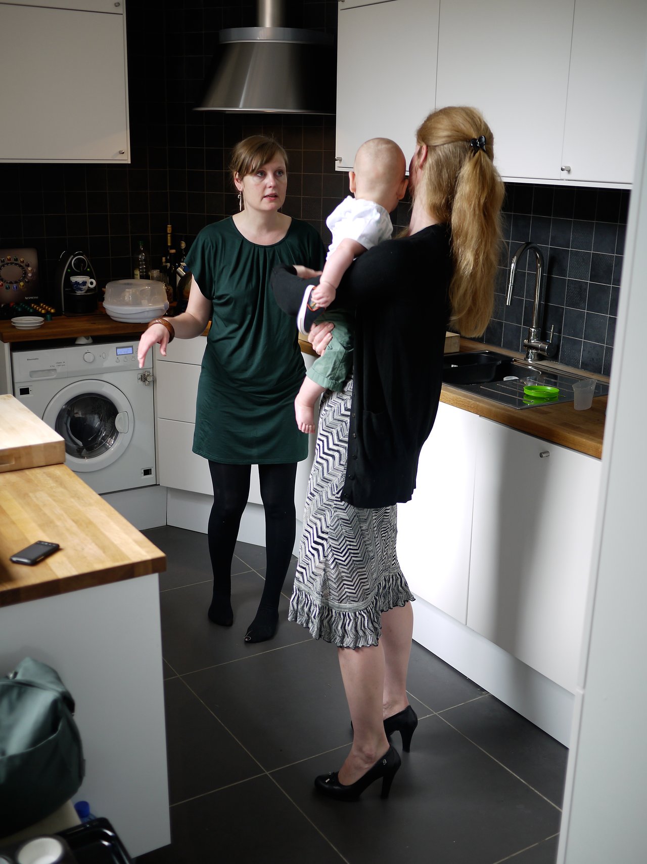 Two women stand in a kitchen, one holding a baby, while they engage in conversation.