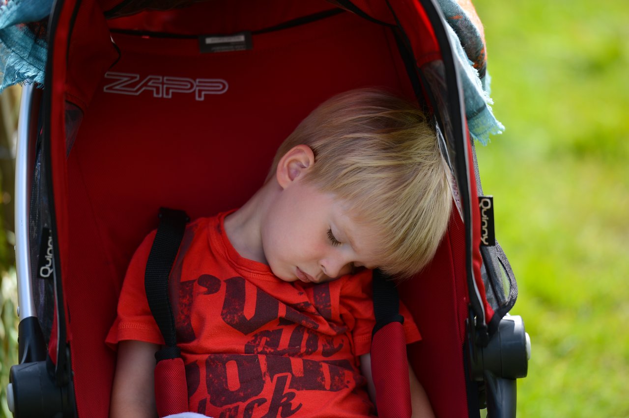 A young child in a red shirt is asleep in a stroller at the playground.