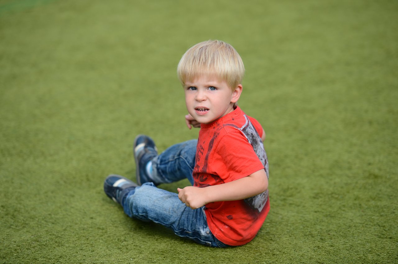 A young child in a red shirt sits on the grass, looking back with a slightly tense expression.