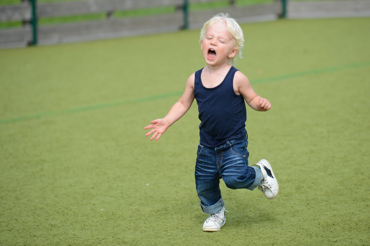 A young child in a navy tank top and jeans runs across a playground, appearing to cry or shout.