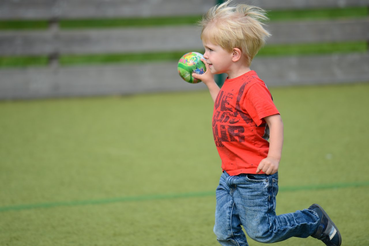 A young child in a red shirt runs across a playground while holding a small colorful ball.