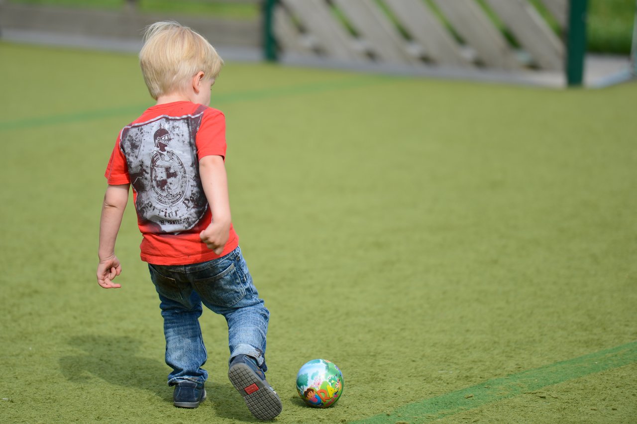 A young child in a red shirt kicks a small colorful ball on a green playground field.