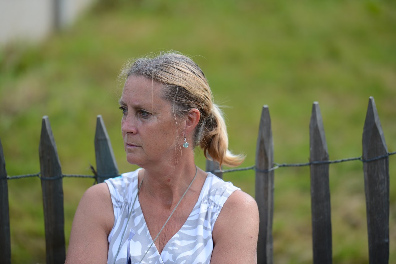 A woman with blonde hair sits near a wooden fence, looking to the side with a neutral expression.
